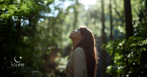 A woman standing in a sunlit forest, head tilted back toward the light