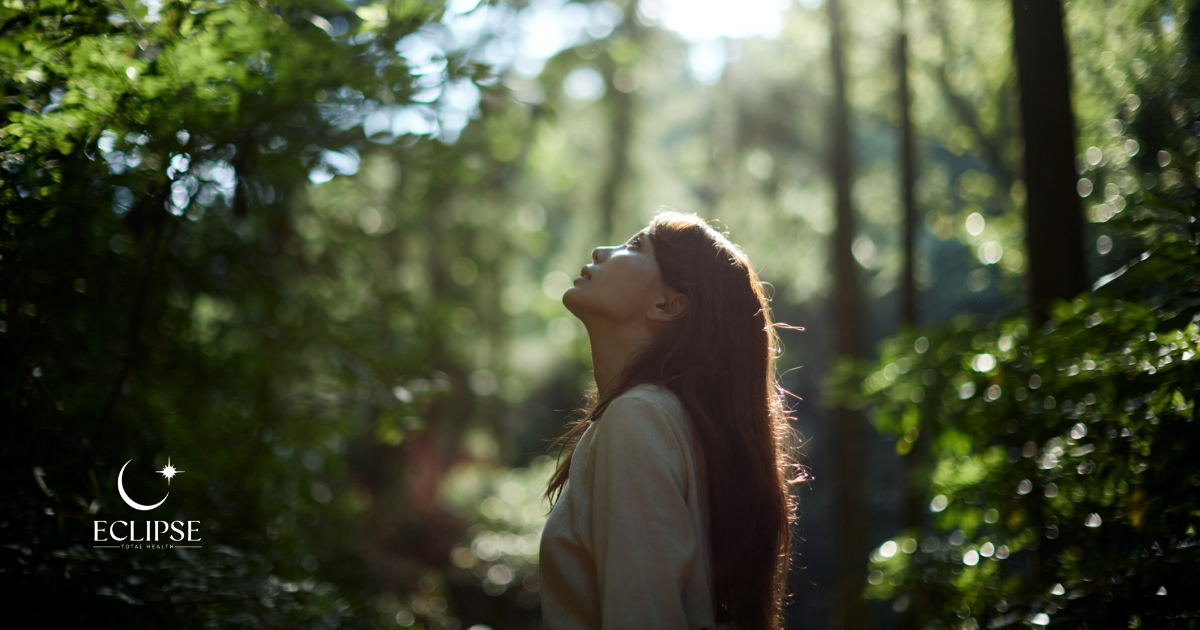 A woman standing in a sunlit forest, head tilted back toward the light
